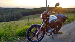 Motocicleta parada em estrada rural ao pôr do sol, com colinas e vegetação no fundo.