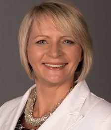 Eine lächelnde Frau mit kurzem, blonden Haar und weißem Blazer vor grauem Hintergrund.