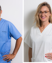 Zwei Personen in medizinischer Kleidung, ein Mann in Blau und eine Frau in Weiß, lächeln.