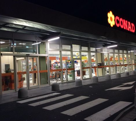 Ingresso di un supermercato Conad illuminato di notte, con strisce pedonali e carrelli visibili.