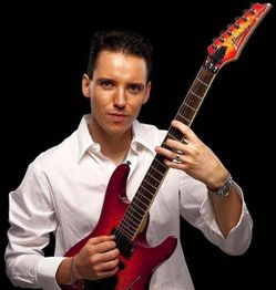 Mann in weißem Hemd hält eine rote Gitarre vor schwarzem Hintergrund.