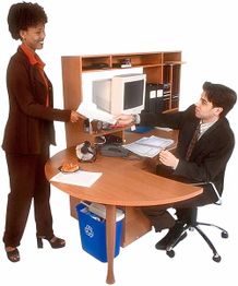 Frau steht am Schreibtisch und reicht Mann ein Papier; Computer und Dokumente sind sichtbar.