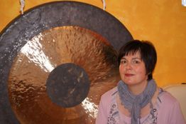 Frau neben einem großen Gong, trägt ein rosa Oberteil und einen grauen Schal, vor gelber Wand.