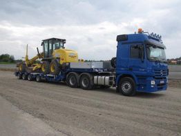 Blauer Lkw transportiert eine gelbe Baumaschine auf einem Anhänger über eine unbefestigte Straße.