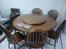 Mesa redonda de madeira com seis cadeiras. Revistas sobre a mesa e outro móvel ao fundo.