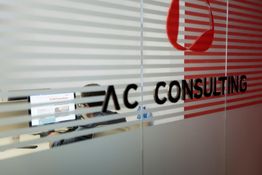 Ufficio con vetro striato e scritta "AC CONSULTING", persona al computer sullo sfondo.