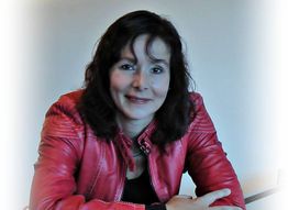 Eine Frau in roter Jacke sitzt lächelnd an einem Tisch, mit neutralem Hintergrund.