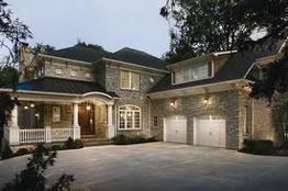 Large stone house with porch and two garages, surrounded by trees, during early evening.