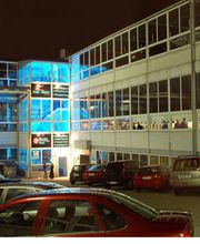 Ein beleuchtetes Gebäude bei Nacht mit Autos auf dem Parkplatz und Vollmond am Himmel.