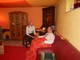 Eine Person auf einem Sofa, in eine Decke gewickelt, und eine stehende Person in einem warmen Raum.