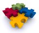 Vier bunte Puzzlestücke in Rot, Blau, Gelb und Grün, ineinandergreifend auf weißem Hintergrund.