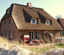 Backsteinhaus mit Reetdach, Strandkorb und Terrasse im Vordergrund, umgeben von Dünen und Gras.