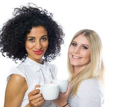 Zwei Frauen mit Kaffeetassen lächeln in die Kamera. Hintergrund ist weiß.