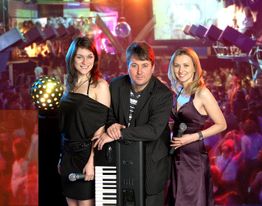 Drei Personen in Abendkleidung vor Keyboard in einem Club mit Disco-Lichtern und Menschenmenge.