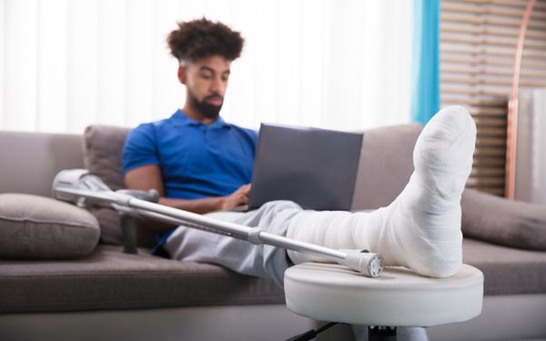 Ein Mann legt sein eingegipstes Bein hoch. Er sitzt auf einer Couch, einen Laptop auf dem Schoß. Neben ihm liegen Krücken.