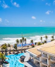 Vista aérea de um resort com piscina à beira-mar, céu azul e praia ao fundo.