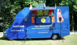 Blauer Puppenspiel-Bus im Grünen mit Bühne, Plakaten und Mikrofonen. "Das Kaspermobil" steht drauf.