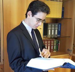 Uomo in giacca esamina un libro aperto in una biblioteca con scaffali di libri dietro di lui.