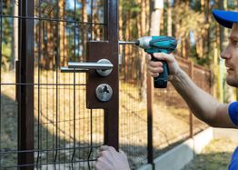 Person fixing a gate lock with a drill in an outdoor setting with trees in the background.