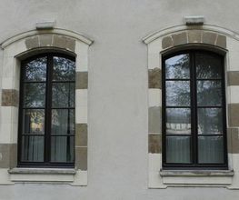 Zwei nebeneinanderliegende Fenster mit braunen Rahmen und gemustertem Mauerwerk.