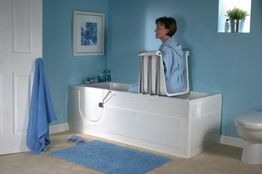 Vrouw in een badlift in een blauw betegelde badkamer, zittend naast een witte badkuip.