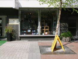 Schaufenster eines Geschäfts mit gelbem Warnschild auf Bürgersteig, Baum rechts daneben.