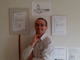 Eine Person lächelnd vor einer Wand mit Zertifikaten und einem „LifeCare Training“-Logo.