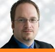 Ein Mann mit Brille und Bart trägt einen Anzug und schaut in die Kamera.
