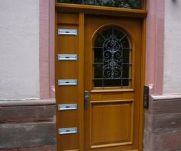 Holztür mit Glaseinsatz, vier Briefkästen links außen in einem rosa umrahmten Gebäude.