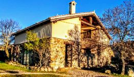 Casa rústica de piedra y madera rodeada de árboles con cielo azul claro.
