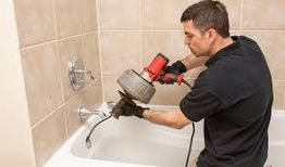 Person using a plumbing snake to unclog a bathtub drain, wearing black gloves and a black shirt.