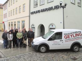 Gruppe posiert vor "Geyling Immobilien" Gebäude mit Firmenfahrzeug im Vordergrund bei Regenwetter.