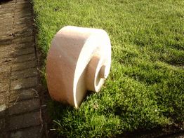 Skulptur einer Schnecke auf grünem Gras neben einem gepflasterten Weg im Sonnenlicht.