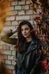 Woman in a black leather jacket poses against a brick wall with autumn leaves.