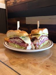 Sandwich halved on a plate with turkey, ham, lettuce, tomato, cheese, and pickles, secured by sticks.