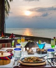 Cena con cócteles frente al mar al atardecer, rodeada de palmeras.