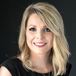 Smiling person with shoulder-length blonde hair, wearing a black top against a dark background.