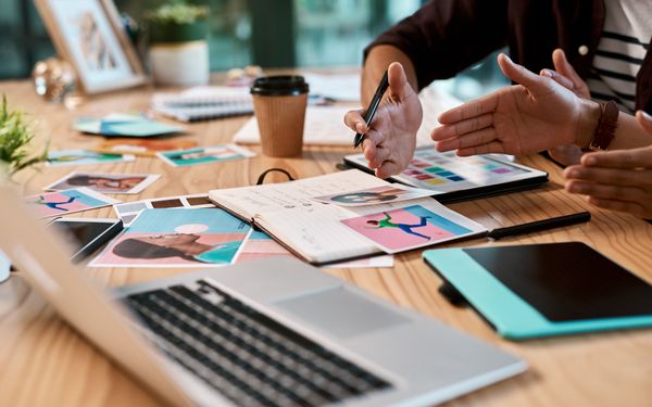 Zwei Personen besprechen gemeinsam einen Businessplan an einem Schreibtisch mit Laptop, Notizbuch, Tablet und Bildmaterial.