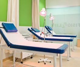 Camillas en una sala de fisioterapia con luces y cortinas, paredes verdes y blancas.