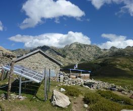 Casa de piedra con paneles solares en las montañas bajo un cielo azul con nubes esponjosas.