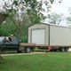 Truck towing a small building on a trailer, with a dog in the foreground on a grassy area.