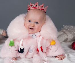 Niedliches Baby in rosa Mantel und Krone, lächelt, umgeben von Kuscheltieren.