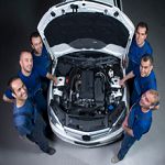 Group of mechanics in blue shirts posing around a car with an open hood, viewed from above.
