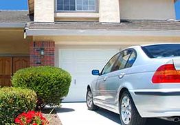 Silver car parked in front of a garage with shrubs and a wooden door nearby.