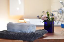 Zusammengerolltes Handtuch auf grauem Tuch, daneben Vase mit Blumen auf einem Holztisch im Badezimmer.