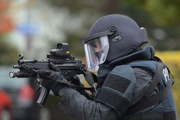 Polizist in Schutzkleidung zielt mit Gewehr, Hintergrund unscharf.