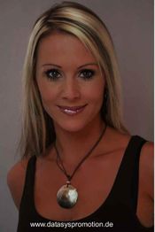 Eine lächelnde Frau mit langem blondem Haar und einem runden Anhänger, vor grauem Hintergrund.