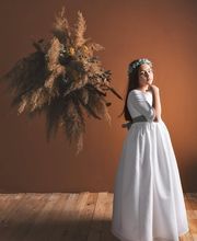 Niña vestida de blanco con corona de flores, junto a un ramo seco en una habitación de madera.