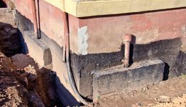 Building foundation with exposed pipes and fresh waterproofing on a sunny day.