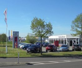 Autohaus mit parkenden Autos, zwei Fahnenmasten und Bäumen darunter, bei sonnigem Wetter.